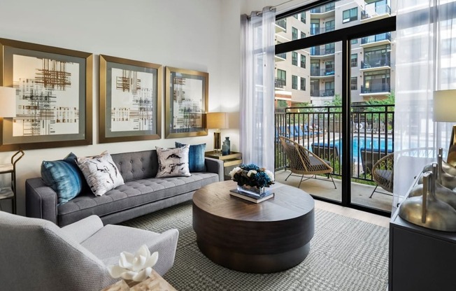 A living room with a grey couch, a coffee table, and a view of a pool and buildings outside.