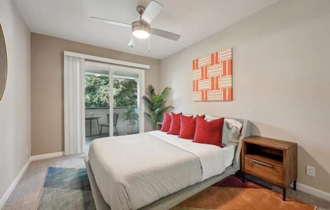 One-Bedroom Apartments in Fremont, CA - Northgate Savoy - Bedroom with Carpeted Flooring, Bed, Wooden Nightstand, Houseplant, Ceiling Fan, Beige Walls, Framed Mounted Mirror, and Sliding Glass Door to Balcony.