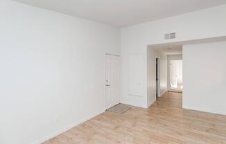 A spacious, empty room with light-colored walls and wooden flooring. There is a closed door on the left, a small welcome mat in front of it, and an entrance to another room visible in the background. The overall atmosphere is bright and clean, suggesting a newly renovated space.