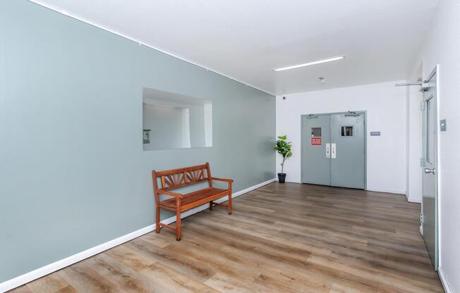 A spacious, well-lit hallway featuring a light gray wall and wooden flooring. There is a simple wooden bench against the wall and a potted plant nearby. Double doors are visible at the end of the hallway, leading to another area, and a large mirror is mounted on the wall.