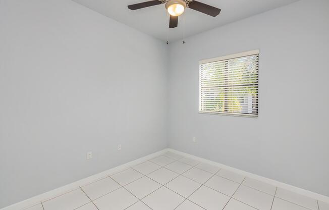 Empty room with light gray walls and white tile flooring. A ceiling fan is mounted in the center of the ceiling. There is a window with blinds on the right side, allowing natural light to enter. The space is clean and uncluttered, suitable for various uses.