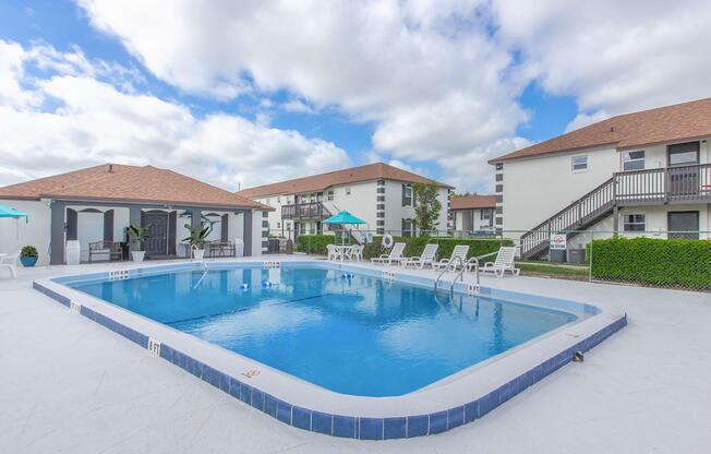 A large swimming pool surrounded by lounge chairs, with umbrellas for shade. In the background, two buildings with a light-colored facade and brown roofs can be seen under a partly cloudy sky. The pool area has a clean, inviting look, perfect for relaxation and recreation.