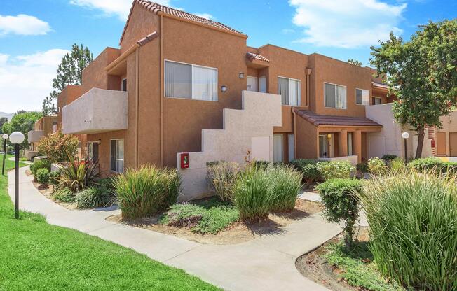 A well-maintained residential complex featuring two-story buildings with a tan exterior. The pathway is surrounded by lush green grass and landscaped shrubs. There are light posts along the walkway, and the sky is clear with a few clouds, creating a bright and inviting atmosphere.