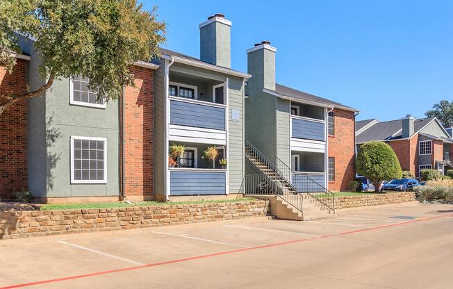 Exterior view of a multi-unit apartment building featuring a mix of brick and painted siding. The building has two levels with balconies adorned with plants. Stairs lead to the upper level. A paved parking area is in front, with greenery in the surrounding landscape and a clear blue sky overhead.