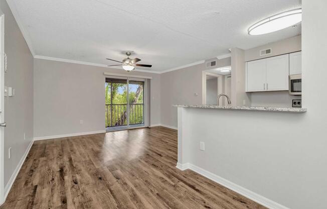 Interior view of a modern apartment featuring an open living space with wood-style flooring, a ceiling fan, and neutral-colored walls. There is a kitchen with white cabinetry and granite countertops visible in the foreground, and a sliding glass door leading to a balcony in the background, surrounded by greenery.