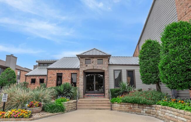 A modern brick and stone house with a metal roof, featuring landscaped flower beds and neatly trimmed bushes. The entrance has a set of double doors and steps leading up to it, with a wide driveway and vibrant flowers enhancing the curb appeal. Clear blue sky in the background.