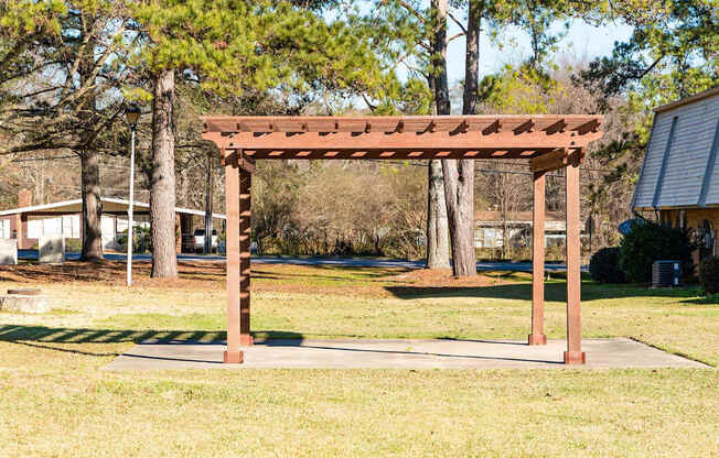 A wooden pergola stands in a grassy area with trees in the background.