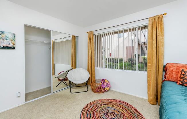 Light and airy bedroom featuring a bed with colorful pillows, a round woven rug on the carpeted floor, a large mirror closet, and a cozy white chair. Natural light streams in through the window, adorned with sheer curtains, offering a view of the outdoors and enhancing the room's inviting ambiance.