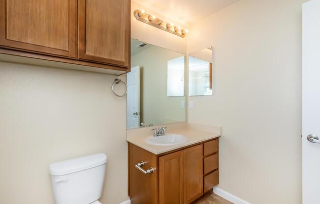 A well-lit bathroom featuring a wooden cabinet above a sink, a mirror with lights, a white toilet, and beige walls. The sink area includes a faucet and is accompanied by wooden drawers underneath. A door is visible in the background.