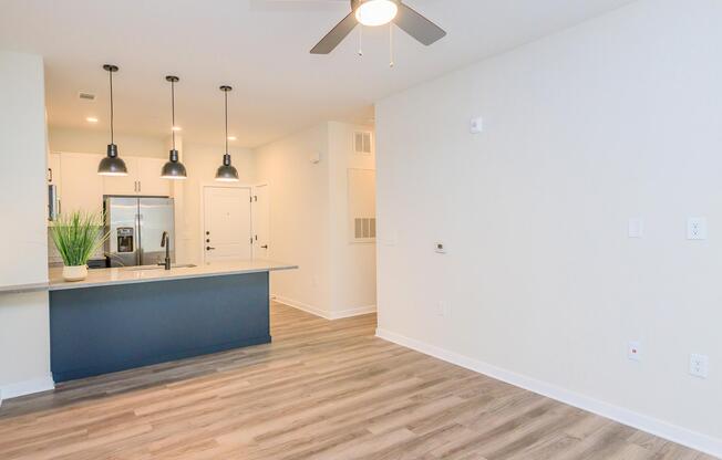 An interior view of a modern living space featuring light-colored walls and hardwood flooring. The kitchen area includes a countertop with bar seating, pendant lights, and stainless steel appliances. A ceiling fan is visible, and there is a decorative plant on the countertop. The space appears bright and spacious.