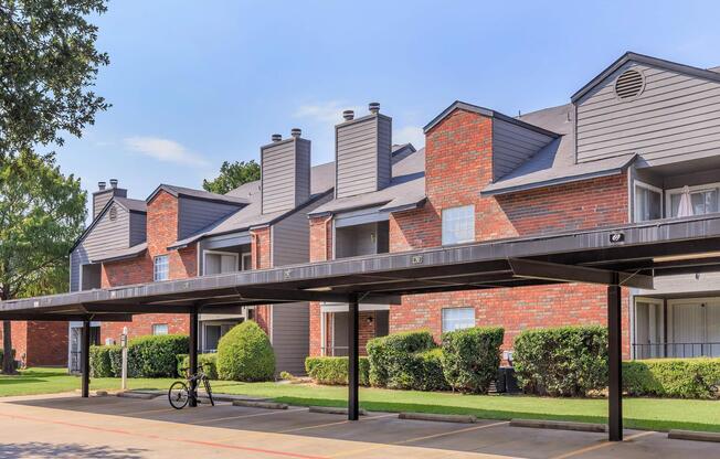 A view of a multi-level apartment complex featuring brick exteriors, sloped roofs, and multiple chimneys. The building has covered parking spaces in the foreground, with neatly trimmed shrubs and grassy areas. Blue sky and trees are visible in the background.
