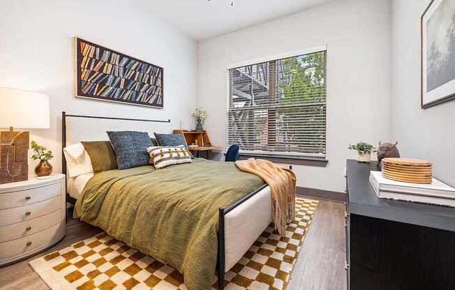 A bedroom with a bed, a dresser, a window with blinds, a painting on the wall, a plant on the nightstand, and a rug on the floor.