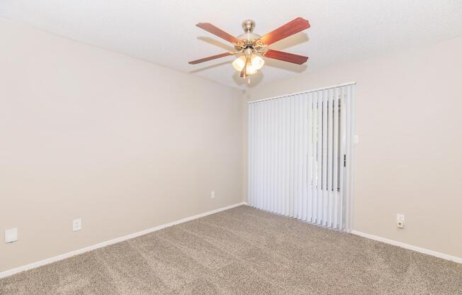 A spacious, unfurnished room with light beige walls and a ceiling fan with wooden blades. The floor is covered in plush carpet, and there is a sliding glass door with vertical blinds leading outside. Natural light streams in through the door.