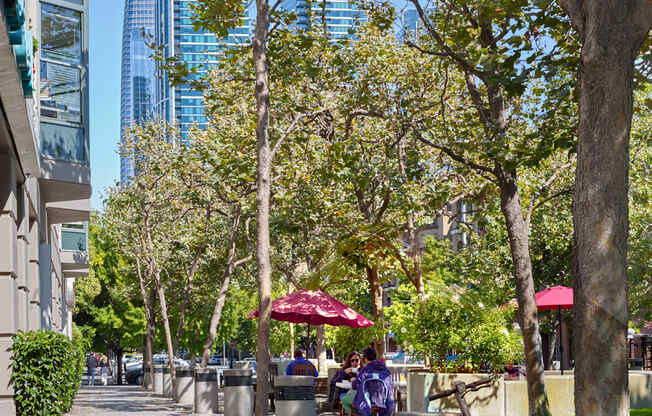 A tree-lined sidewalk with people sitting at tables under umbrellas.