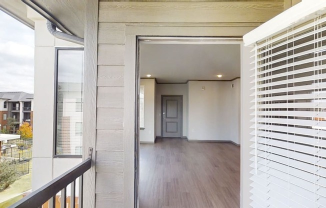 View from a balcony here at 49 West Apartments into an empty apartment with wood flooring, neutral walls, and a closed door. Nearby buildings and trees are visible outside.
