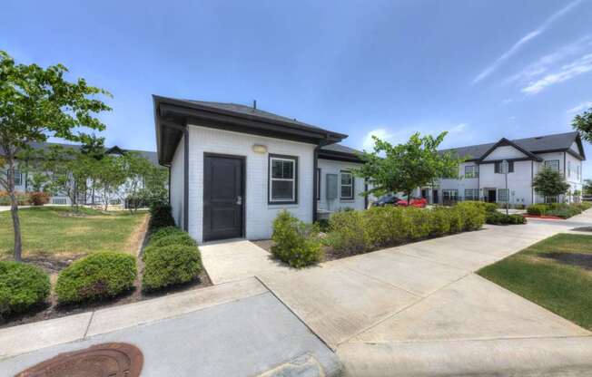 A small white house with a black door and windows is surrounded by a well-maintained lawn and a sidewalk.