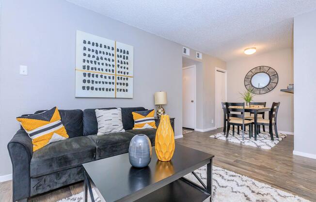 A modern living room featuring a dark sectional sofa with decorative pillows, a coffee table with a decorative vase, and a bright yellow lamp. The space has grey walls and a textured rug, with a dining area visible in the background, including a round table and chairs. Natural light filters in from the nearby doorways.