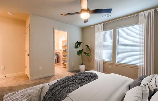 Raymore, MO Townhomes - Bedroom With Ceiling Fan, Windows, Large Walk-In Closet, Carpet Flooring, And Two Windows.