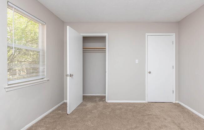 A vacant room with light gray walls and beige carpet. It features a window with blinds on the left, an open closet with a single shelf on the back wall, and two doors—one leading to the closet and a second white door on the right. Natural light fills the space, creating a bright and airy atmosphere.