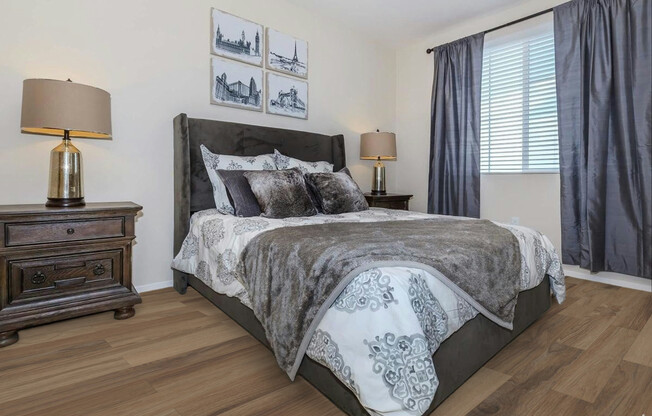 A cozy bedroom featuring a queen-sized bed with decorative pillows and a soft gray blanket. There are two bedside lamps on ornate wooden nightstands, and the walls are adorned with framed artwork. Natural light filters through a window with gray curtains, and the flooring is made of light wood.
