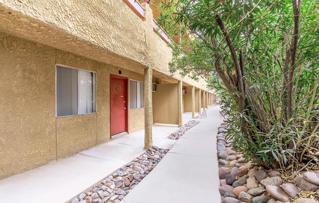 A pathway runs alongside a series of beige-colored buildings with red doors. Lush greenery, including bamboo-like plants, lines one side of the walkway. The scene depicts a well-maintained outdoor area of a residential or commercial building complex.