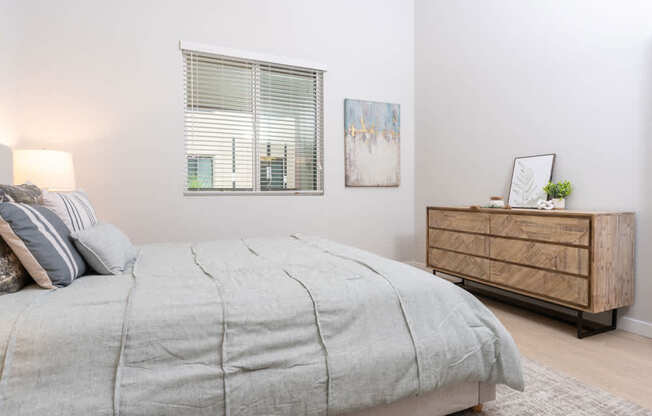 A bed with a grey comforter and pillows sits in a room with a wooden dresser and a painting on the wall.