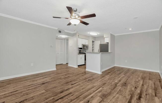 A spacious, modern living area featuring a ceiling fan, hardwood-style flooring, and light gray walls. The open layout connects to a kitchen with stainless steel appliances and white cabinetry. A sliding door leads to a small outdoor space, providing natural light and a welcoming atmosphere.