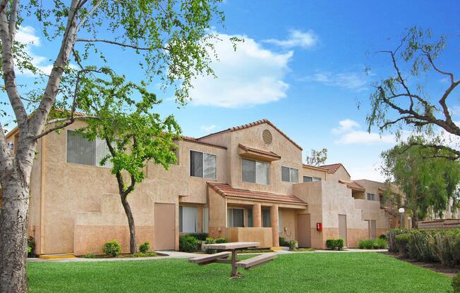 A view of a residential complex with beige stucco buildings set against a bright blue sky. Lush green grass surrounds the buildings, with a few trees and shrubs present. A picnic table can be seen in the foreground, suggesting an inviting outdoor space for residents.