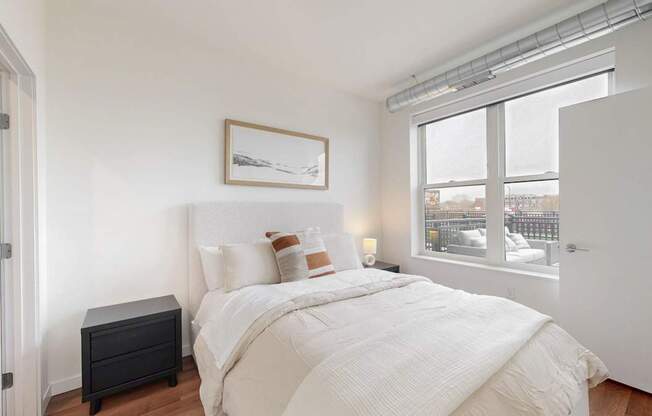 A bedroom with a large bed, a nightstand, and a window overlooking a cityscape.