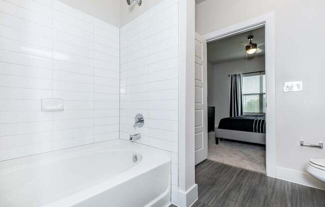 A white bathroom with a tub and a bedroom in the background.