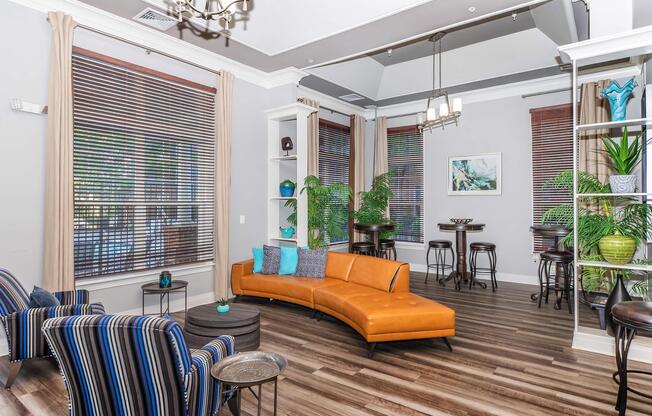 A modern living room featuring a curved orange couch, two striped chairs, and small tables. Large windows with wooden blinds allow natural light in, complemented by potted plants. Shelves display decorative items, and there is a bar area with stools in the background, creating a stylish and inviting atmosphere.