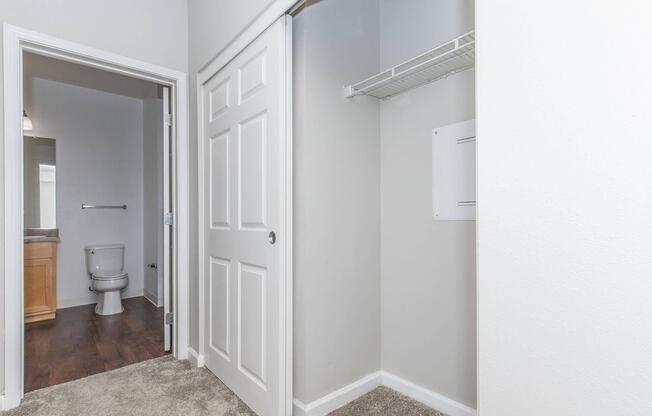 Interior view of a bathroom area featuring a partially open door to a closet on the left, leading to a toilet and sink visible in the background. The walls are painted in a light color, and the floor is covered with carpet. A small shelf is mounted in the closet, and there is a closet door on the right.