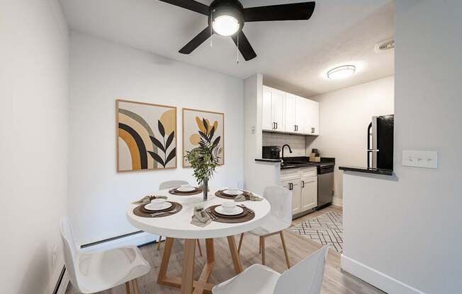 A dining room with a white table and chairs, a ceiling fan, and a kitchen area in the background.