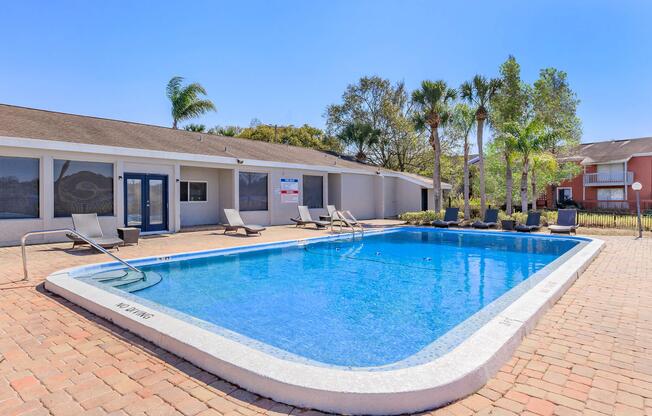 A clear blue swimming pool surrounded by lounge chairs, with palm trees in the background. The pool area is sunny and inviting, featuring a brick patio. A building with large windows is visible beside the pool, along with a for-rent sign.