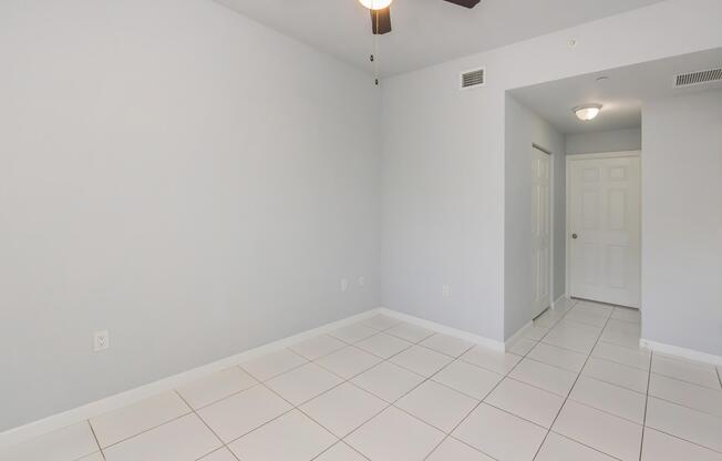 Empty room with light gray walls and white tiled flooring. A ceiling fan hangs from the ceiling. There is a doorway leading to another space on the right and a light fixture on the ceiling. The room appears spacious and well-lit, ready for furnishing or decoration.