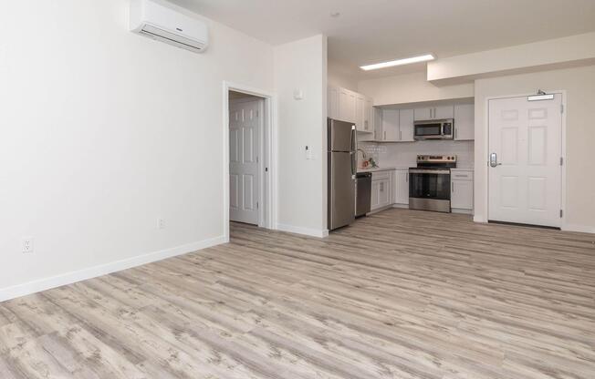 Interior view of a modern apartment featuring an open layout with light wood flooring. The kitchen includes stainless steel appliances and white cabinetry. A doorway leads to another room, and there is an air conditioning unit mounted on the wall. The walls are painted in a light color, enhancing the spacious feel of the area.