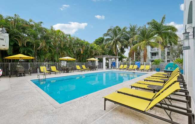 A pool with yellow sun loungers and umbrellas surrounded by palm trees.