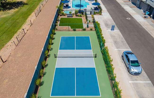 A tennis court is surrounded by a green fence and a parking lot.