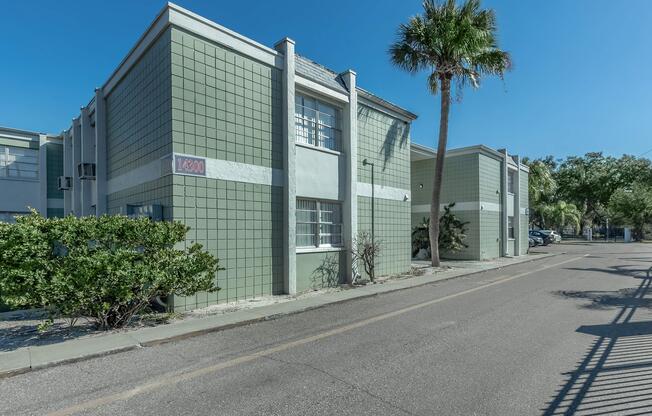 A low-rise building with a green and white exterior, featuring large windows. The scene includes a palm tree nearby and a tree bush lined along the street. The image captures a clear blue sky and a quiet street with parked cars in the background.