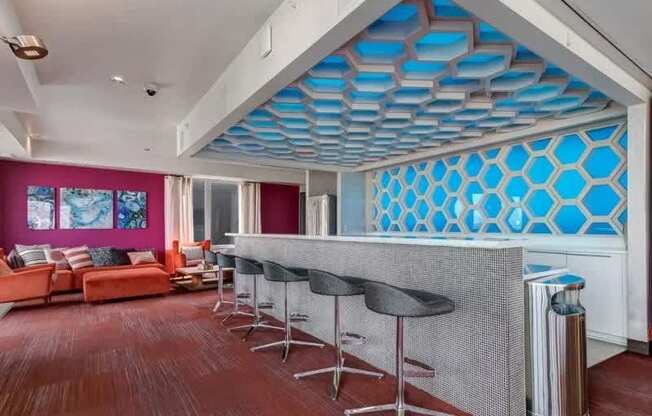 A stylish resident lounge bar here at Blue and Lime showcasing a long tiled counter with modern barstools, a striking blue geometric wall feature, and a matching patterned ceiling detail. Warm wood flooring leads to a cozy seating area with a coral sofa, accent chairs, and colorful artwork, creating a vibrant and social atmosphere.