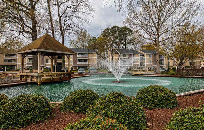 A gazebo sits in the middle of a pond with a fountain in the foreground.