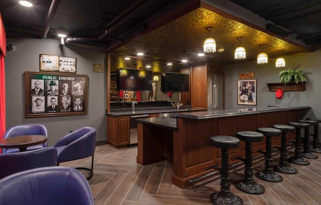 A stylish home bar featuring dark wood cabinets, a black countertop, and high-backed stools. The walls are adorned with framed historical photos and decorative elements, while warm lighting fixtures provide an inviting ambiance. Cozy seating is arranged nearby, enhancing the bar's social atmosphere.