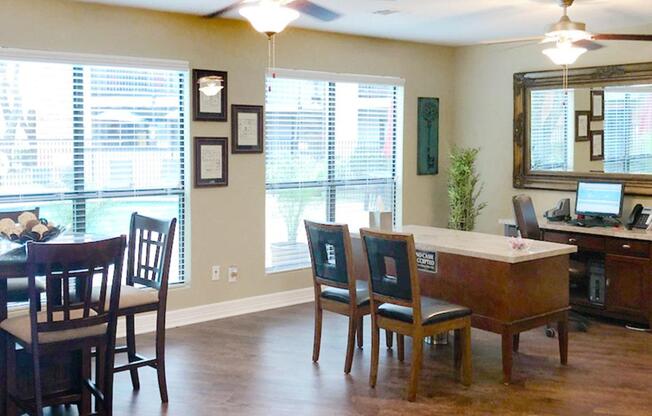 An interior view of a modern office or reception area featuring a wooden desk, several seating options including chairs and a high-top table, and large windows with blinds. The decor includes framed documents and plants, creating a welcoming atmosphere.