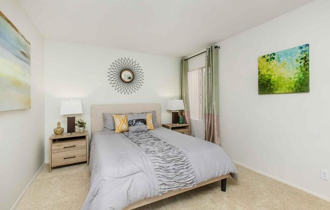A cozy bedroom featuring a neatly made bed with gray bedding and decorative pillows. There are two bedside tables with lamps on either side, and a round mirror above the bed. The walls are adorned with two framed artworks, and the room has light-colored carpet and large windows with sheer curtains.