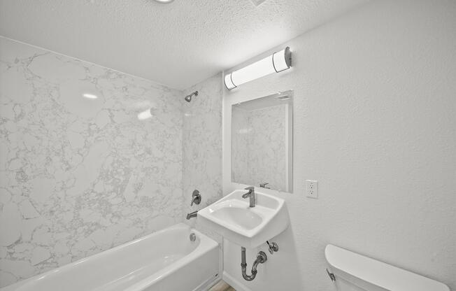 A modern bathroom featuring a white bathtub, a wall-mounted sink, and a large mirror. The walls are adorned with a light-colored marbled pattern, and there is a bright ceiling light above. A clean, minimalist design is highlighted by light wood flooring.