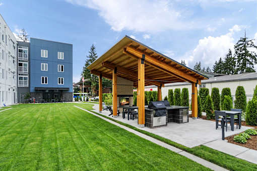 A wooden pavilion is surrounded by a grassy area and a patio with a grill.