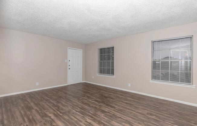 An empty room with light beige walls and a textured ceiling. It features two windows with blinds, a white door, and a floor made of dark wooden planks. The space is well-lit, appearing spacious and ready for furnishing.