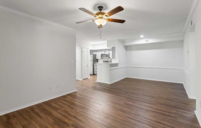 A spacious, light-colored living area with a ceiling fan and hardwood flooring. To the left, there is a small kitchen area visible with modern appliances. White walls create a bright atmosphere, and a half wall separates the living space from the kitchen. Natural light illuminates the room.