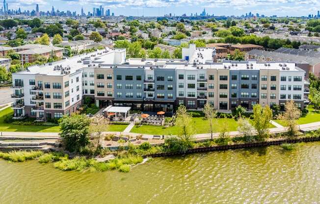 A large apartment complex sits on the edge of a body of water with a city skyline in the background.