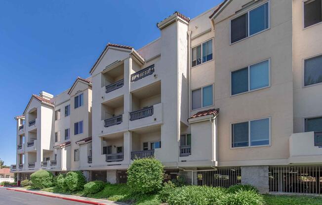 A row of modern apartment buildings with beige exteriors and terracotta roofs. The structure features multiple balconies, large windows, and surrounding green shrubs. The sky is clear and blue, indicating a sunny day.